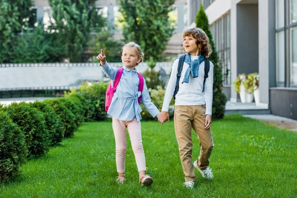 Cute little schoolkids with backpacks holding hands and looking away while walking on green lawn — Stock Photo