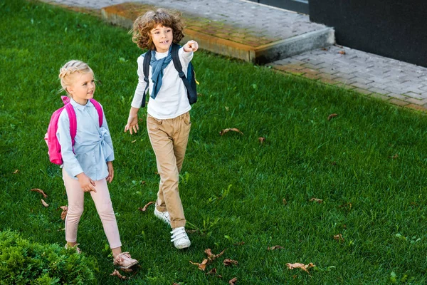 Visão de alto ângulo de escolares bonitos com mochilas andando no gramado verde e apontando para longe com o dedo — Fotografia de Stock