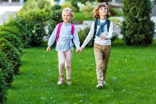 Visão comprimento total de adoráveis estudantes felizes com mochilas de mãos dadas e andando no gramado verde — Fotografia de Stock