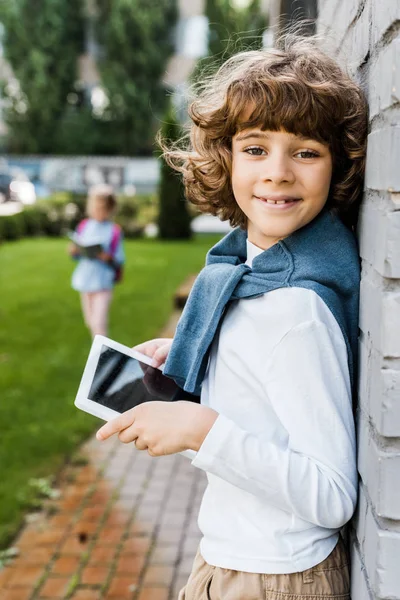 Adorable escolar utilizando tableta digital y sonriendo a la cámara - foto de stock