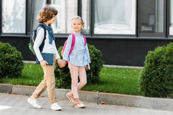 Niedliche kleine Schulkinder mit Rucksäcken, die Händchen halten und auf der Straße laufen — Stockfoto