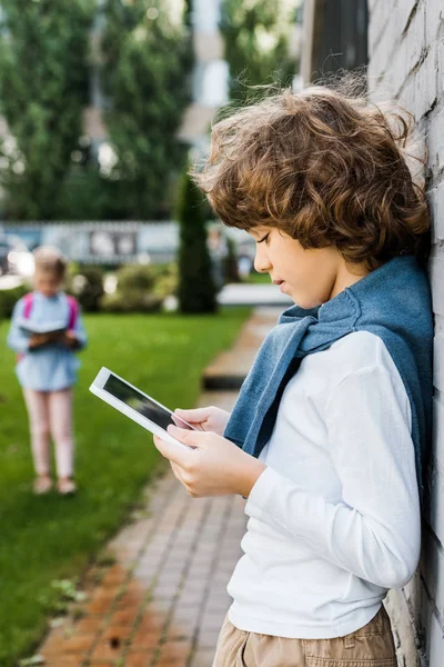 Side view of cute preteen schoolboy leaning at brick wall and using digital tablet — Stock Photo