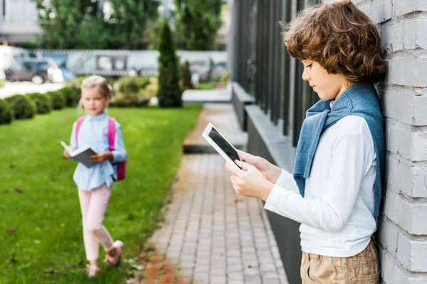 Вид сбоку школьника с цифровым планшетом и школьницы, держащей книгу снаружи — стоковое фото