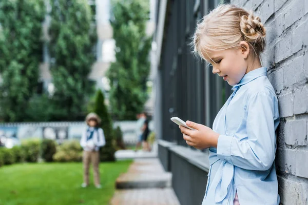 Vue latérale de mignon enfant souriant penché au mur de briques et en utilisant un smartphone — Photo de stock