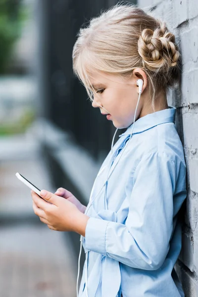 Vista lateral de niño adorable en auriculares usando tableta digital y apoyado en la pared de ladrillo - foto de stock