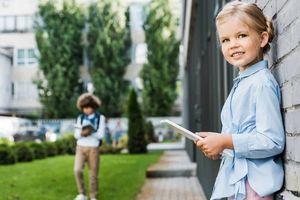 Cute little schoolgirl using digital tablet and smiling at camera while leaning at brick wall — Stock Photo