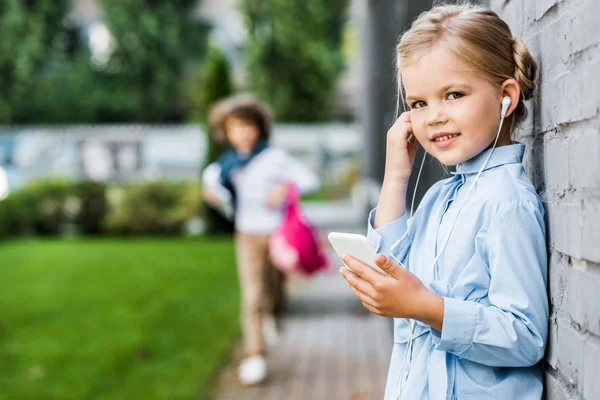 Cute little child in earphones using smartphone and smiling at camera — Stock Photo