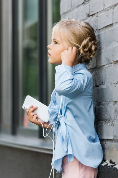 Vista lateral de un niño adorable en auriculares que sostienen el teléfono inteligente, apoyado en la pared de ladrillo y mirando hacia otro lado - foto de stock