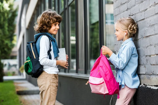 Cute little schoolchildren with backpacks smiling each other outside — Stock Photo