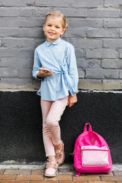 Cute little schoolgirl using smartphone and smiling at camera while leaning at brick wall with pink backpack — Stock Photo