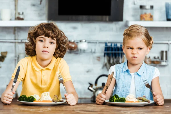 Mignons enfants malheureux tenant des couverts et regardant la caméra tout en étant assis à table et en mangeant des légumes — Photo de stock