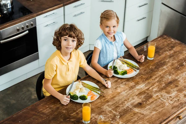 Vista de ángulo alto de lindos niños felices comiendo verduras saludables y sonriendo a la cámara - foto de stock