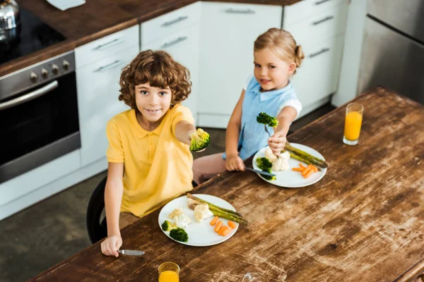 Vista ad alto angolo dei bambini che tengono le forchette con broccoli e sorridono alla fotocamera — Foto stock