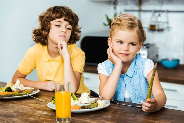 Bambini annoiati a guardare gli asparagi seduti a tavola con le verdure sui piatti — Foto stock