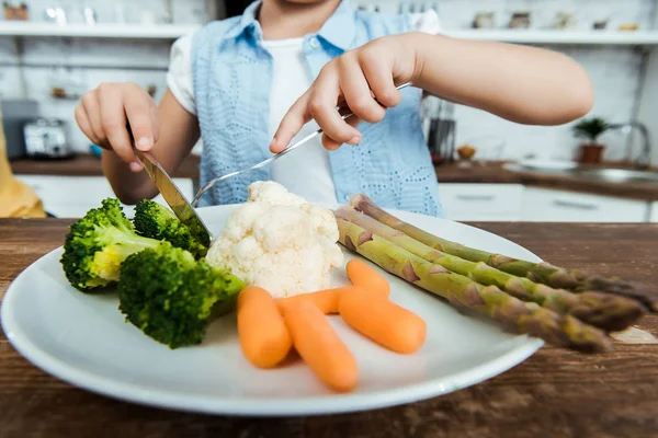 Обрезанный снимок ребенка, держащего вилку с ножом и питающегося здоровыми овощами — стоковое фото