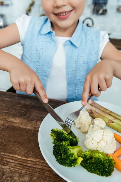 Plan recadré d'un enfant souriant tenant une fourchette avec un couteau et mangeant du brocoli — Photo de stock