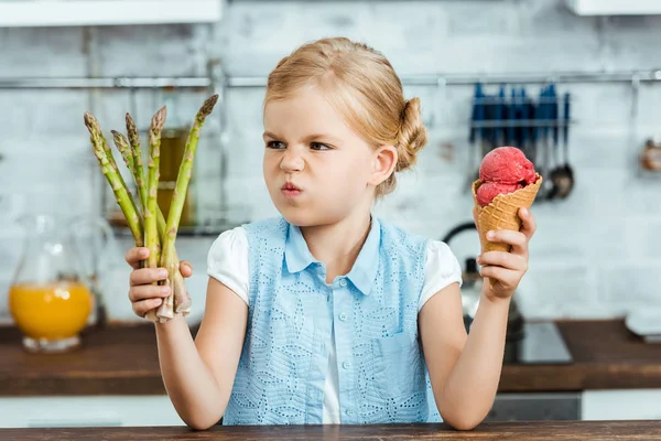 Infeliz niño sosteniendo delicioso cono de helado y espárragos saludables - foto de stock