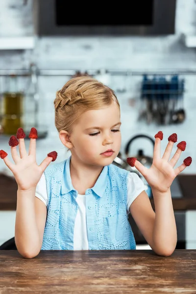 Lindo niño pequeño sentado en la mesa y mirando a las frambuesas frescas maduras en los dedos - foto de stock