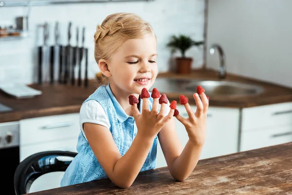 Niedliches glückliches Kind sitzt am Tisch und schaut auf reife frische Himbeeren an den Fingern — Stockfoto