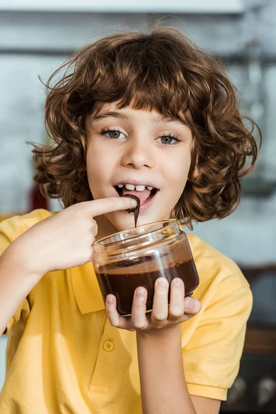 Entzückendes glückliches Kind isst Schokoladenaufstrich aus Container und lächelt in die Kamera — Stockfoto