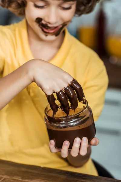 Schnappschuss eines glücklichen Kindes, das Schokoladenaufstrich mit der Hand aus einem Glas isst — Stockfoto