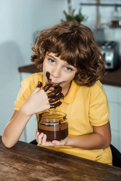 Cute little boy eating sweet chocolate spread from glass jar and looking at camera — Stock Photo