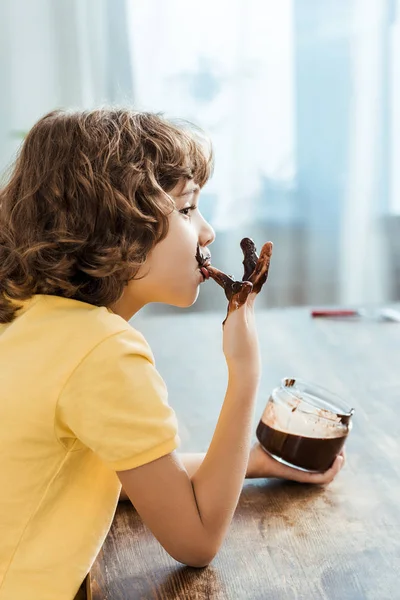 Seitenansicht des niedlichen kleinen Jungen leckt Finger mit leckerem Schokoladenaufstrich — Stockfoto