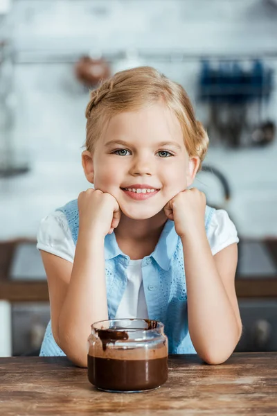 Lindo niño feliz sentado en la mesa con chocolate extendido en frasco y sonriendo a la cámara - foto de stock