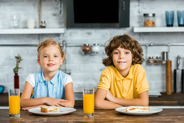 Милые счастливые дети, улыбающиеся в камеру, сидя за столом со стаканами сока и вкусными бутербродами — стоковое фото