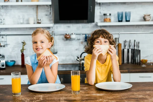 Adorables niños felices comiendo sabrosos sándwiches y sonriendo a la cámara - foto de stock