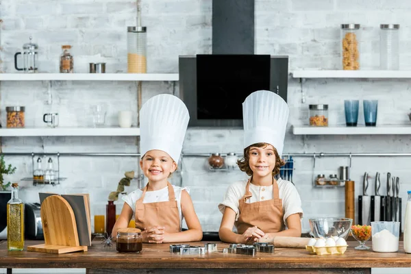 Entzückend glückliche Kinder in Schürzen und Kochmützen lächeln in die Kamera, während sie gemeinsam in der Küche kochen — Stockfoto