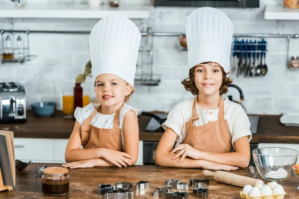 Süße glückliche Kinder in Schürzen und Kochmützen lächeln in die Kamera, während sie gemeinsam in der Küche kochen — Stockfoto
