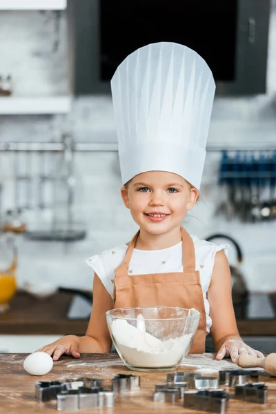 Süßes glückliches Kind in Schürze und Kochmütze, das Teig kocht und in die Kamera lächelt — Stockfoto