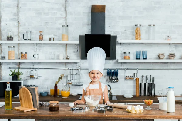 Adorable kid in chef hat and apron cooking dough and smiling at camera — Stock Photo