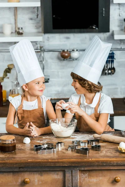 Niños lindos en delantales y sombreros de chef preparando masa para galletas y sonriendo entre sí - foto de stock