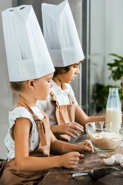 Side view of adorable kids in chef hats and aprons preparing dough for cookies in kitchen — Stock Photo