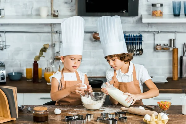 Adorables enfants dans des chapeaux de chef et tabliers préparant la pâte pour les biscuits dans la cuisine — Photo de stock