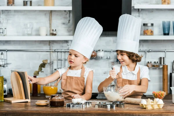 Lindos niños pequeños en sombreros de chef preparando masa y mirando el libro de cocina - foto de stock
