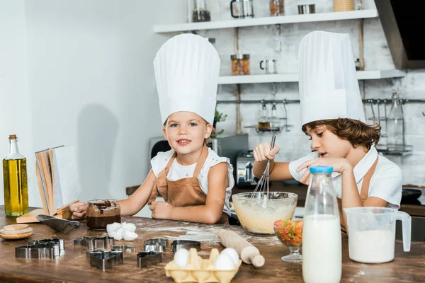 Petits enfants mignons dans des tabliers et des chapeaux de chef préparant la pâte pour les biscuits et souriant à la caméra — Photo de stock