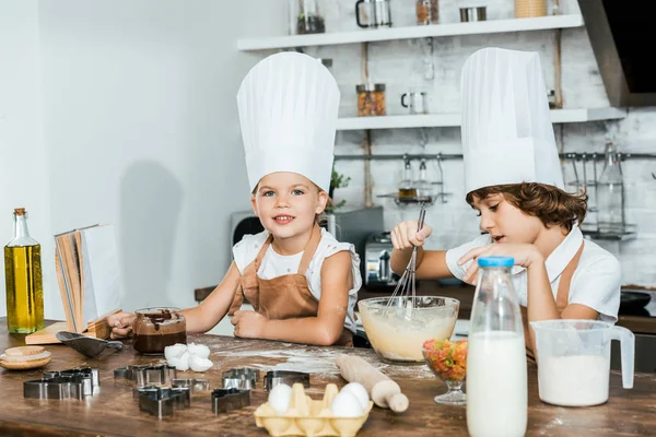 Niedliche kleine Kinder in Schürzen und Kochmützen bereiten Teig für Plätzchen zu und lächeln in die Kamera — Stockfoto