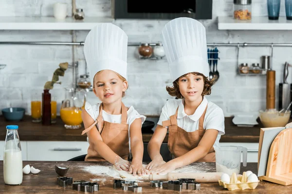 Entzückende Kinder in Schürzen und Kochmützen bereiten gemeinsam Teig für leckere Plätzchen zu — Stockfoto