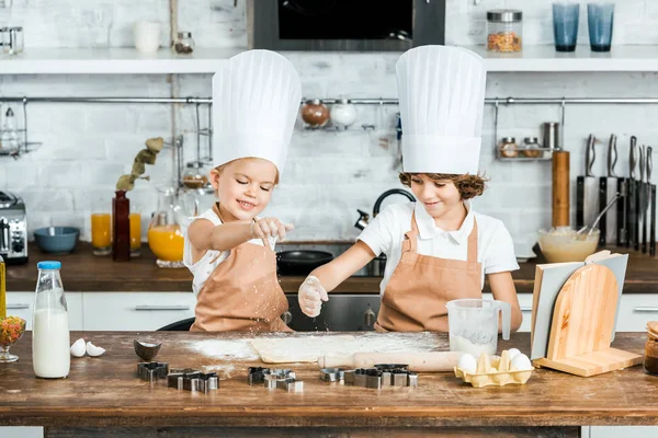 Süße glückliche Kinder in Schürzen und Kochmützen bereiten gemeinsam Teig für leckere Plätzchen zu — Stockfoto