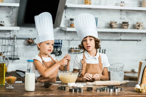 Entzückende Kinder in Schürzen und Kochmützen bereiten gemeinsam Teig für leckere Plätzchen zu — Stockfoto