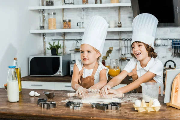 Lindos niños felices en sombreros de chef preparando masa para galletas y mirando hacia otro lado - foto de stock