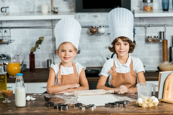 Niedliche glückliche Kinder in Kochmützen bereiten Teig für Plätzchen vor und lächeln in die Kamera — Stockfoto