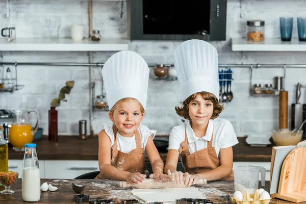 Adorables niños felices en sombreros de chef preparando masa para galletas y sonriendo a la cámara - foto de stock