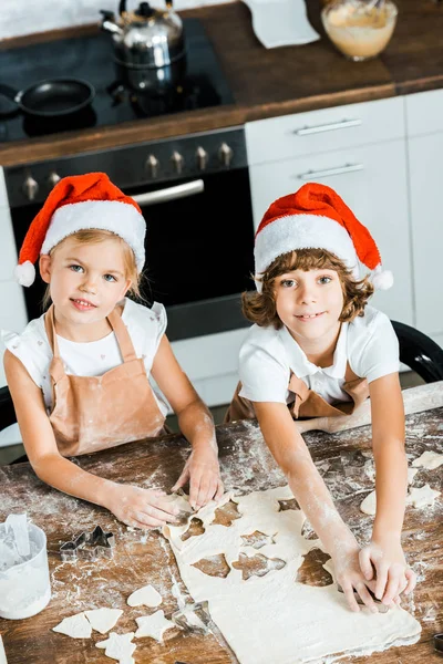 Vue grand angle d'adorables enfants en chapeaux de Père Noël préparant des biscuits de Noël et souriant à la caméra — Photo de stock