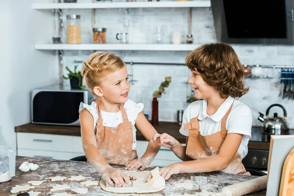 Милые счастливые дети в фартуках готовят вкусное печенье и улыбаются друг другу — стоковое фото