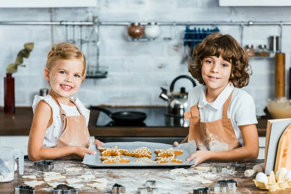 Милые счастливые дети в фартуках, держащие поднос для выпечки с имбирным печеньем и улыбающиеся в камеру — стоковое фото