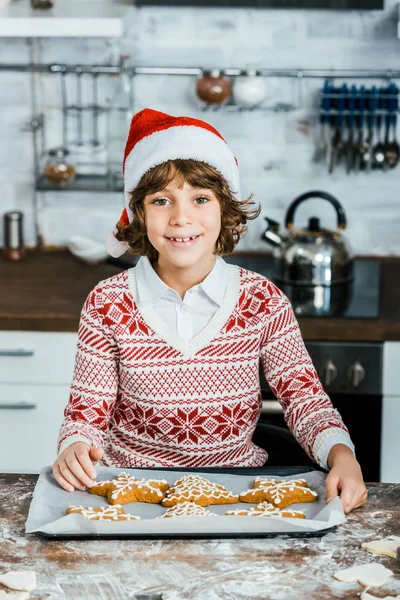 Entzückender glücklicher Junge mit Weihnachtsmütze hält Backblech mit Ingwerplätzchen und lächelt in die Kamera — Stockfoto